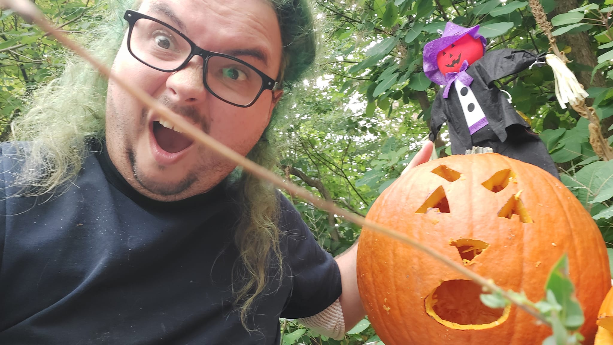 A man with long green hair and no beard has an exadurated surprised expression and holds a large punpkin with a suprrised face carved on it